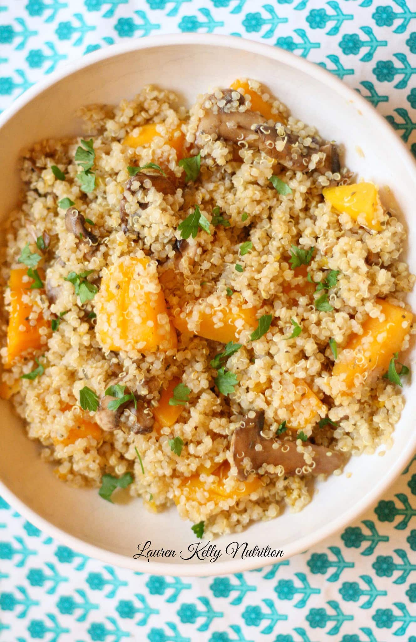 • A white bowl filled with creamy Mushroom and Squash Quinoa Risotto, garnished with fresh parsley and placed on a teal patterned tablecloth.