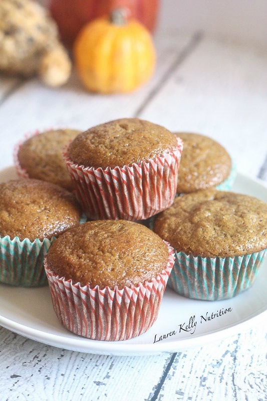 Picture of stacked pumpkin gingerbread muffins, some in red striped liners and some in green striped liners on a white plate on a wooden background with pumpkins in the background.