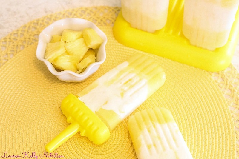 Horizontal picture of two popsicles on  a yellow placemat with a. small white bowl of chopped pineapple in upper left corner and some more popsicles in the upper right corner.
