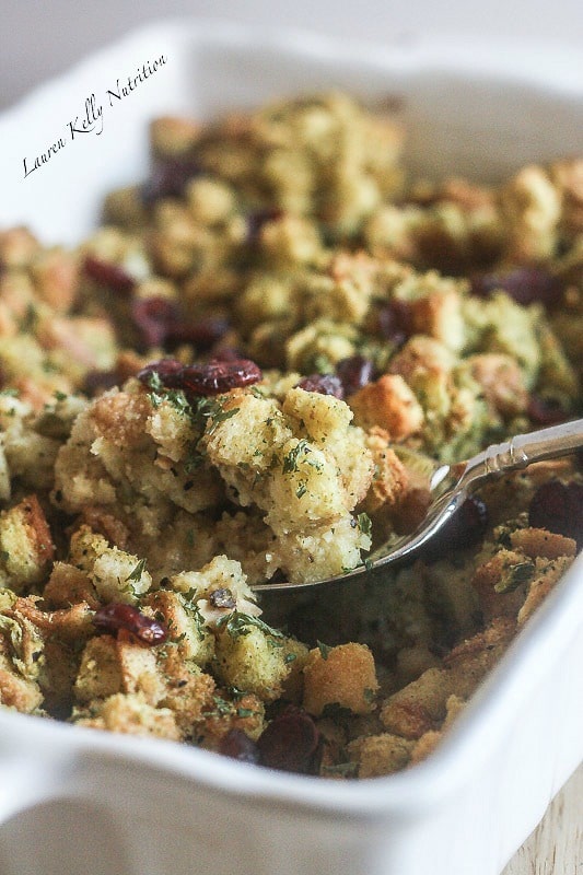 Close up of cornbread stuffing in a white baking dish and a spoonful of stuffing in a silver spoon in front.