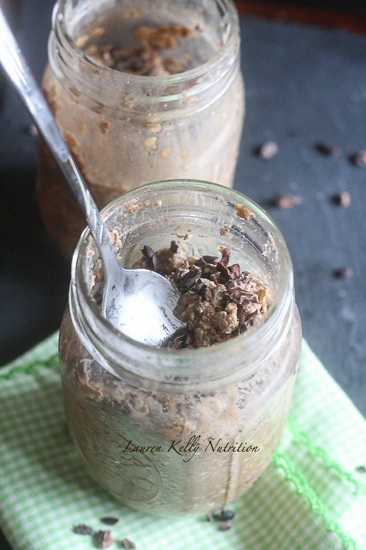 Close up picture of a clear jar of overnight oats with cacao nibs and a silver spoon coming out the top, another jar of oats is in the background.