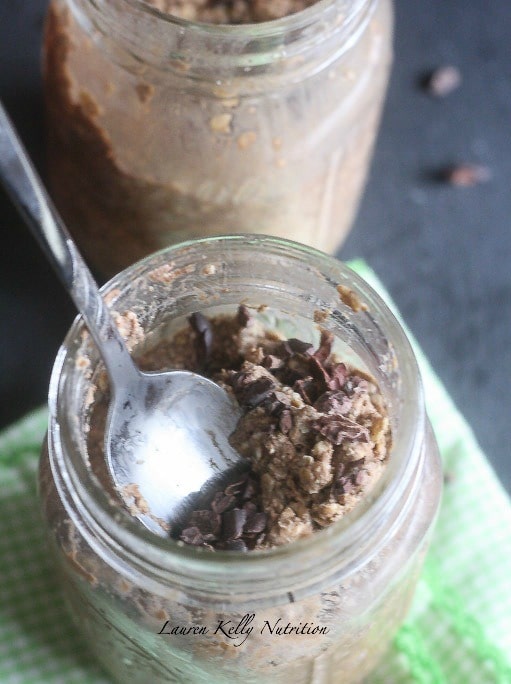 Close up of a clear jar of overnight oats overhead, with cacao nibs and a silver spoon coming out the top.