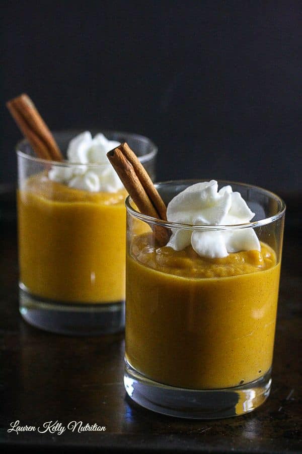 Picture of two glass jars of pumpkin mousse with one cinnamon stick inside and a dollop of whipped cream on top with a dark background.