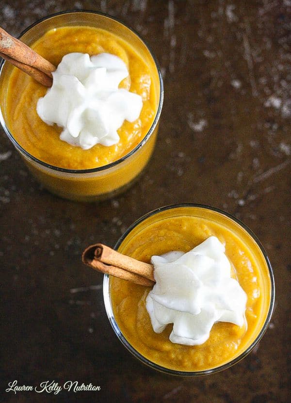 Overhead picture of the tops of two glass jars with pumpkin mousse inside, one cinnamon stick coming out the side of each jar and a dollop of whipped cream on top on a dark background.