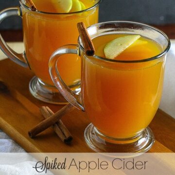 Close up of spiked apple cider in glass mugs with floating apple slices and cinnamon sticks in it.