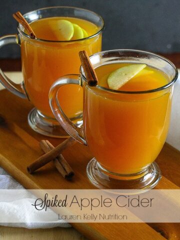 Close up of spiked apple cider in glass mugs with floating apple slices and cinnamon sticks in it.