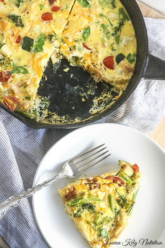 Overhead view of vegetable turkey frittata in a dark skillet  with a piece cut out and on a white plate.