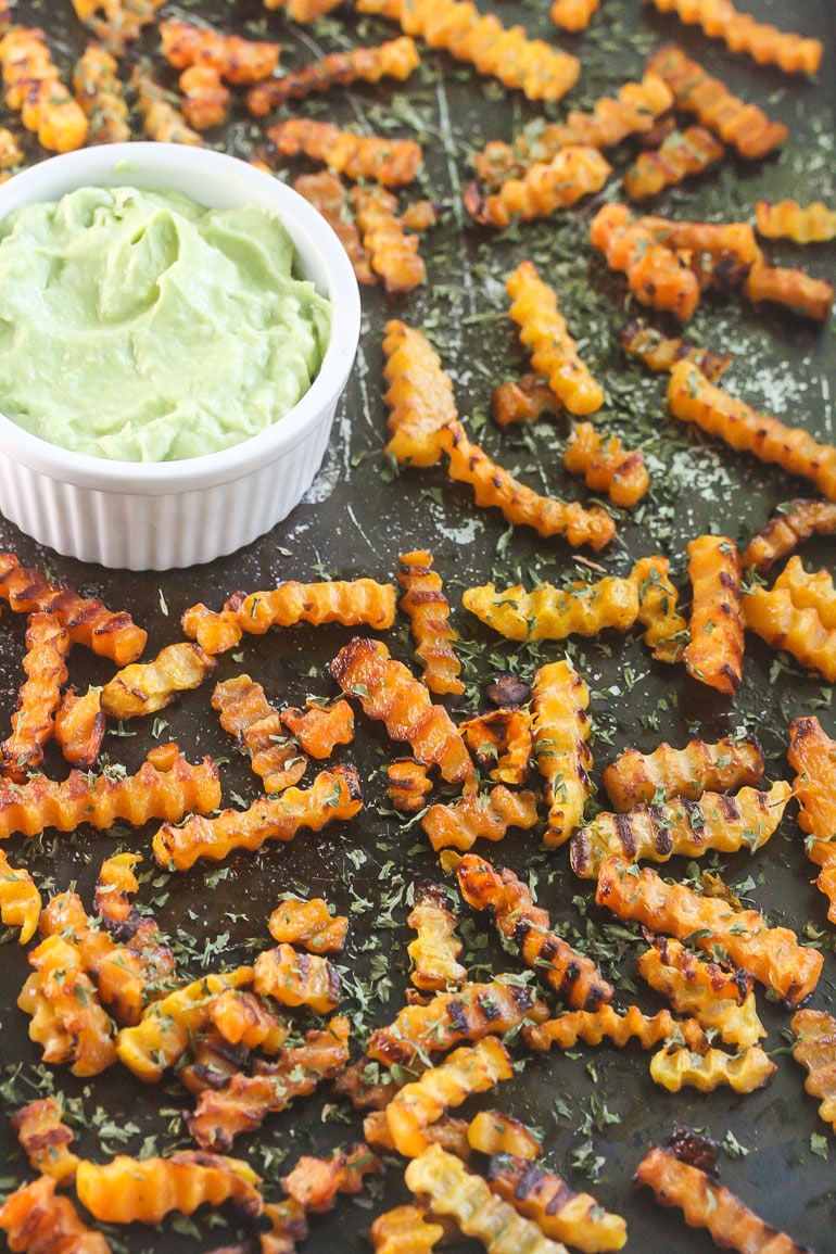 Overhead picture of crinkled butternut squash fries on a dark baking tray scattered with a hand dipping one fry into the small white bowl of avocado dipping sauce.