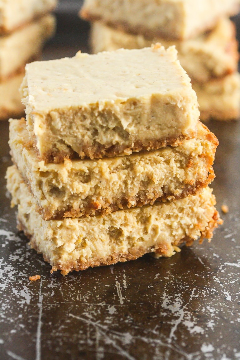 Close up picture of three no bake pumpkin cheesecake bars stacked on top of each other on a dark baking sheet with other pumpkin cheesecake bars behind them.