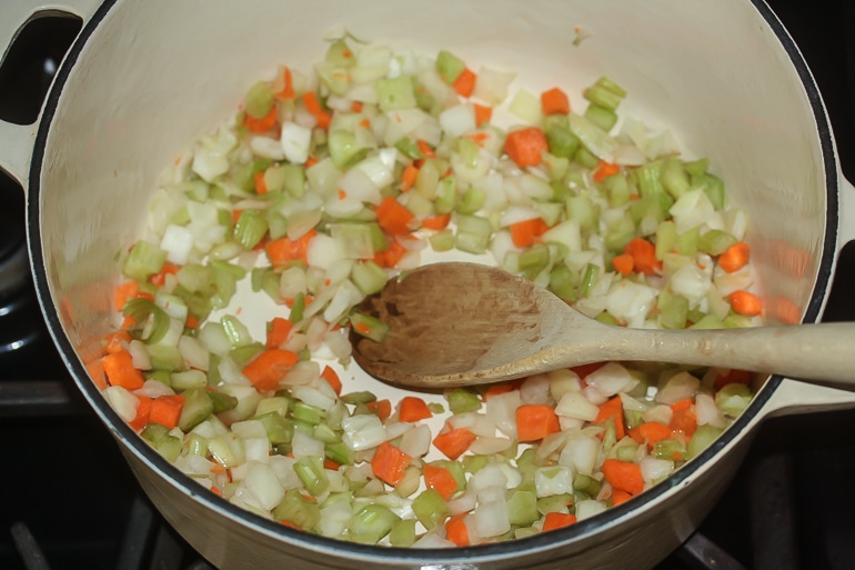 Overhead horizontal picture of diced carrots, celery and onions in an off white dutch oven on a stove with a wooden spoon in it.