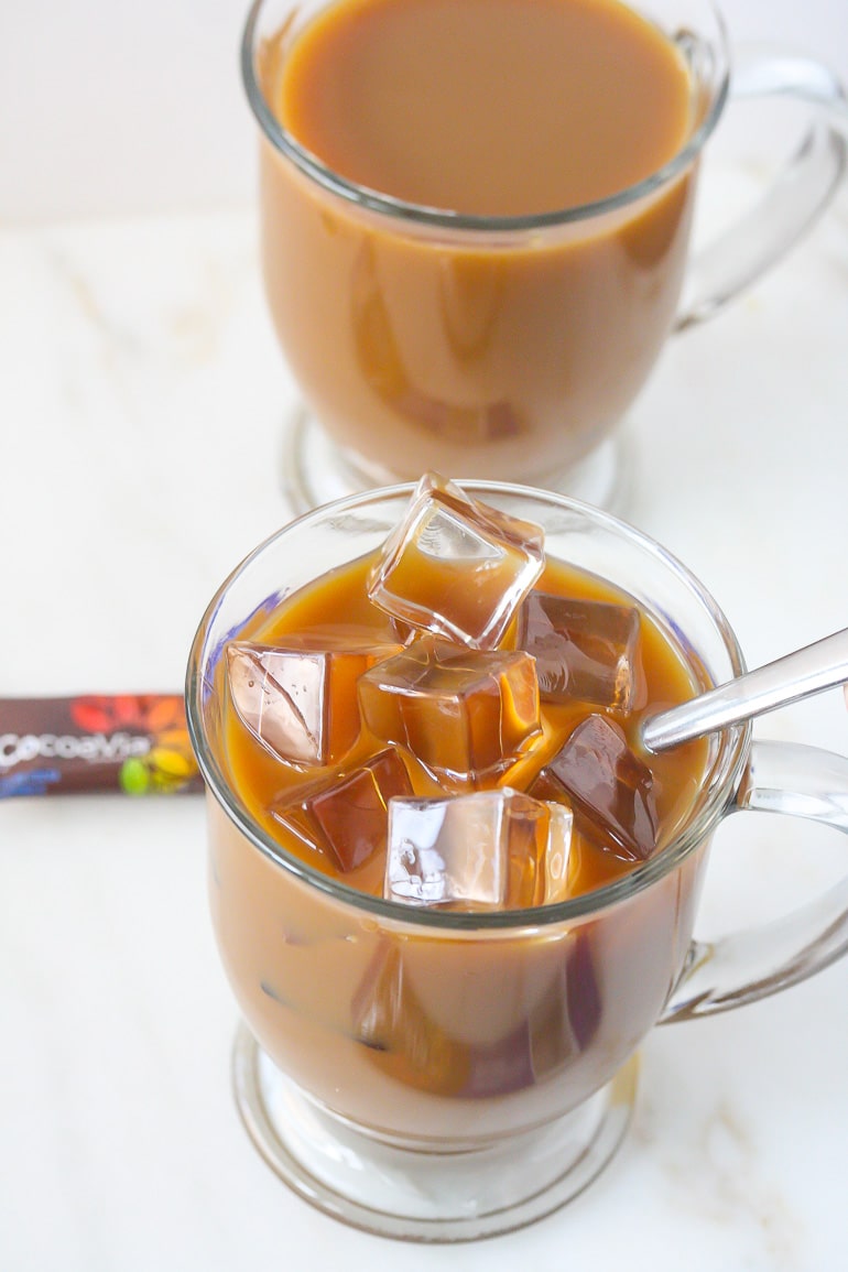 Close up of Iced Mocha Coffee with ice cubes in a clear mug with spoon hanging out and another clear mug with coffee and no ice cubes above it.