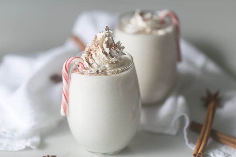 Two glasses of skinny peppermint eggnog with a small candy cane on the side and whipped cream on top.