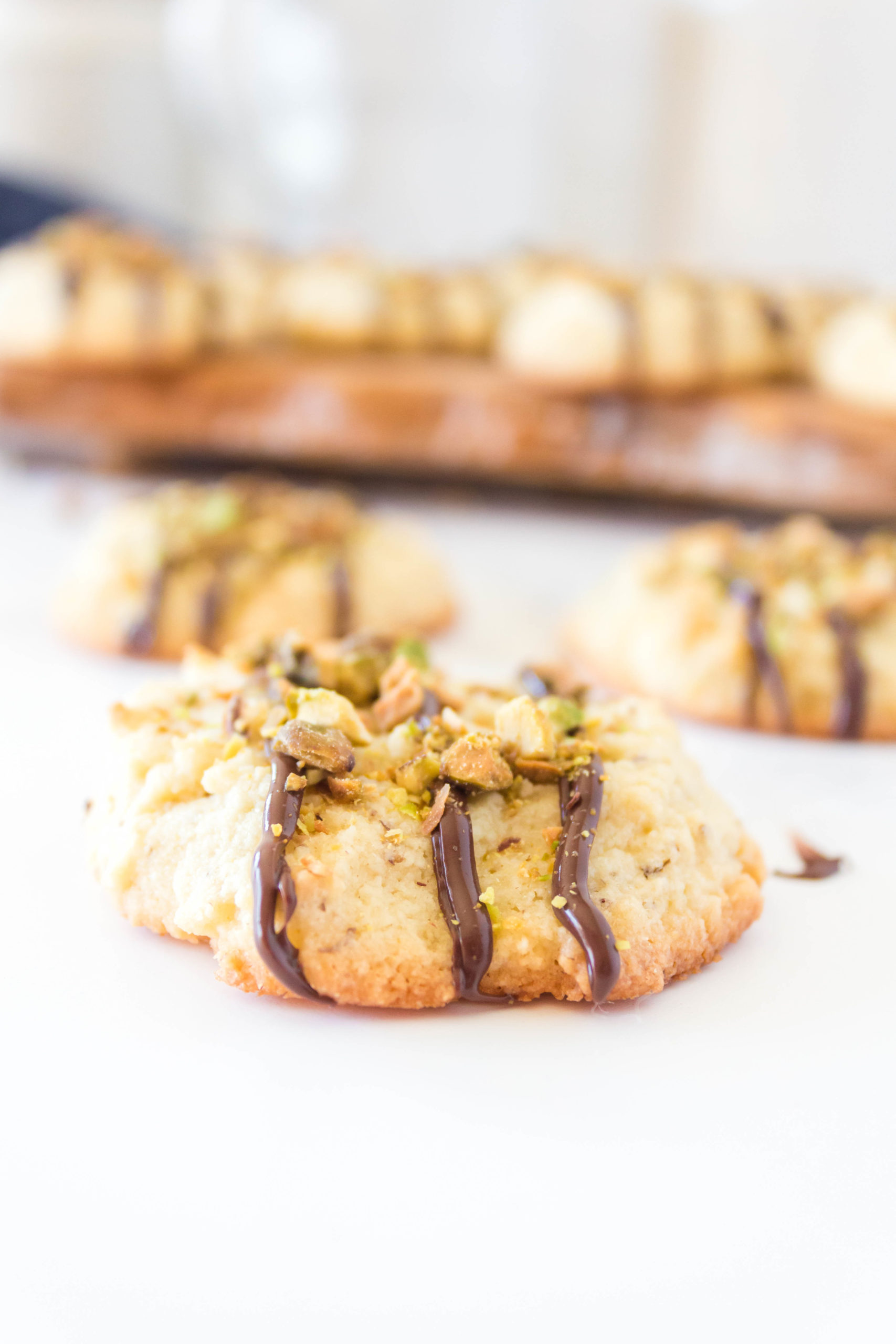 One keto butter cookie with drizzled chocolate and chopped pistachios in top with others in the background.