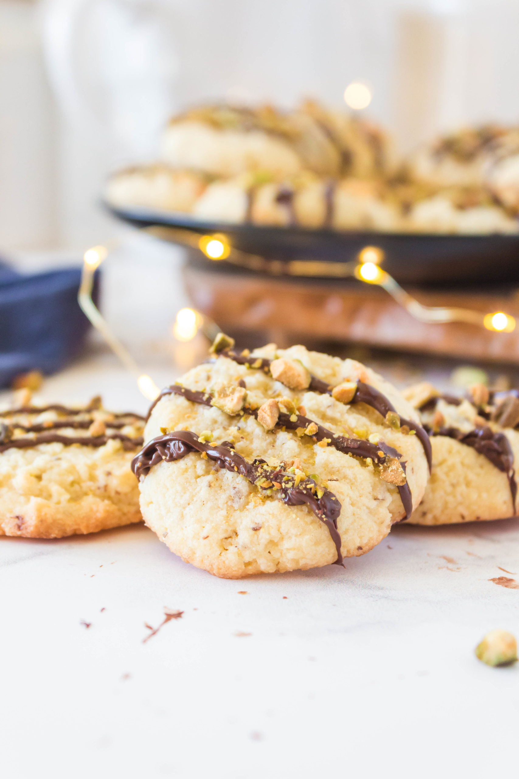 Cookies with chocolate drizzle and chopped pistachios on top with one close up in front.