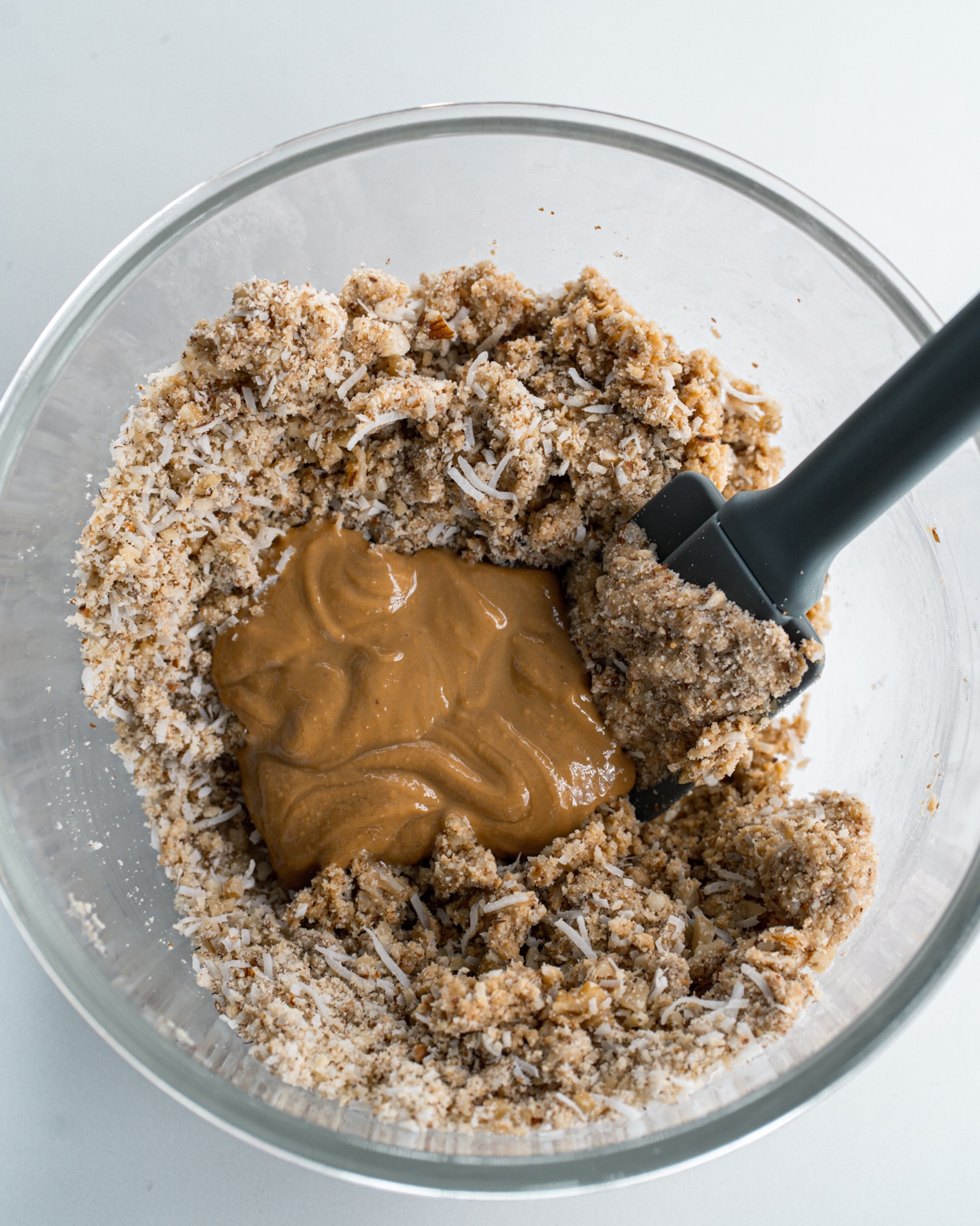 Dry ingredients with almond butter in a glass bowl with grey spatula.