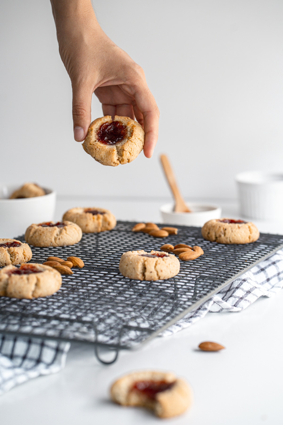 Almond flour thumbprint cookies on a wire rack with almonds scattered on the rack and a hand picking up one cookie.