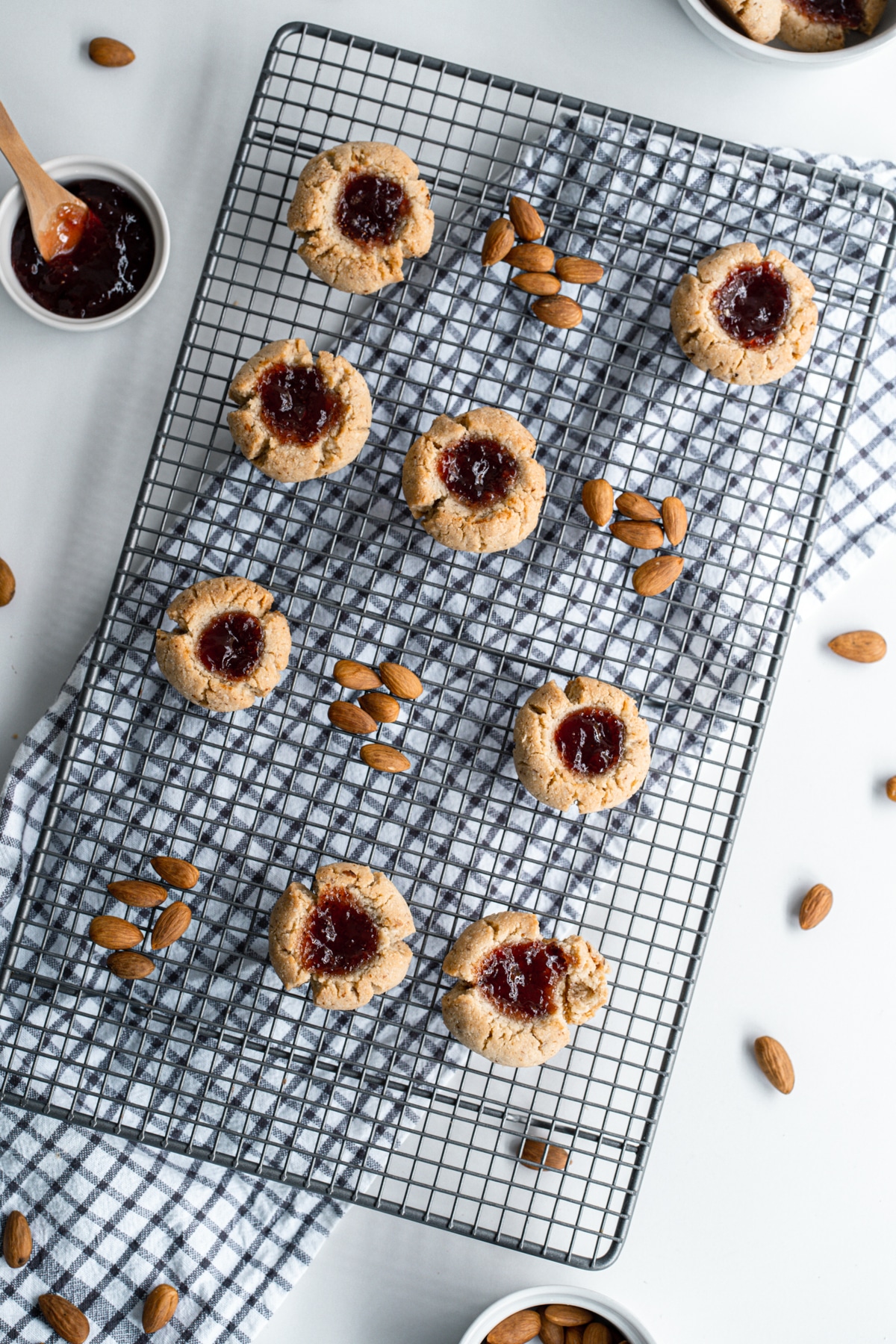 Almond flour thumbprint cookies on a wire rack with scattered almond and a small white bow of strawberry jam in upper left corner.