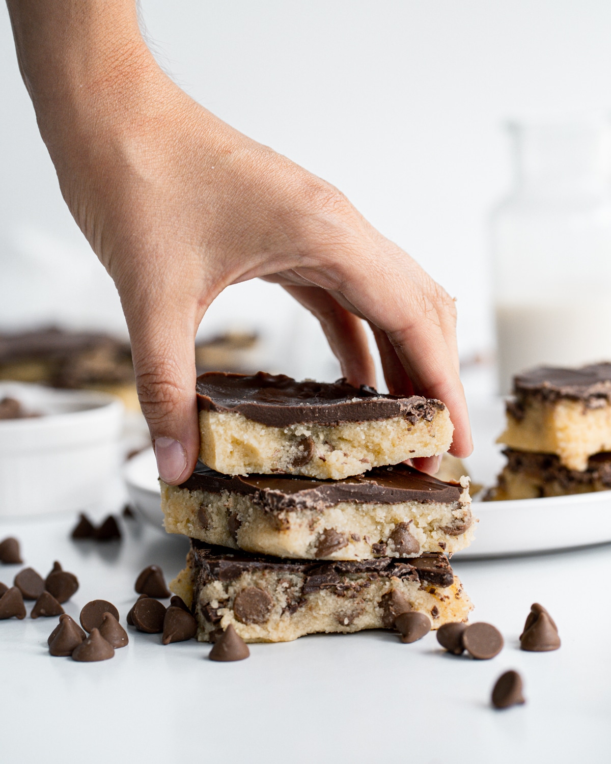 Picture of 3 no bake cookie dough bars stacked on top of each with chocolate chips spread in front and a hand grabbing the bar on top. 