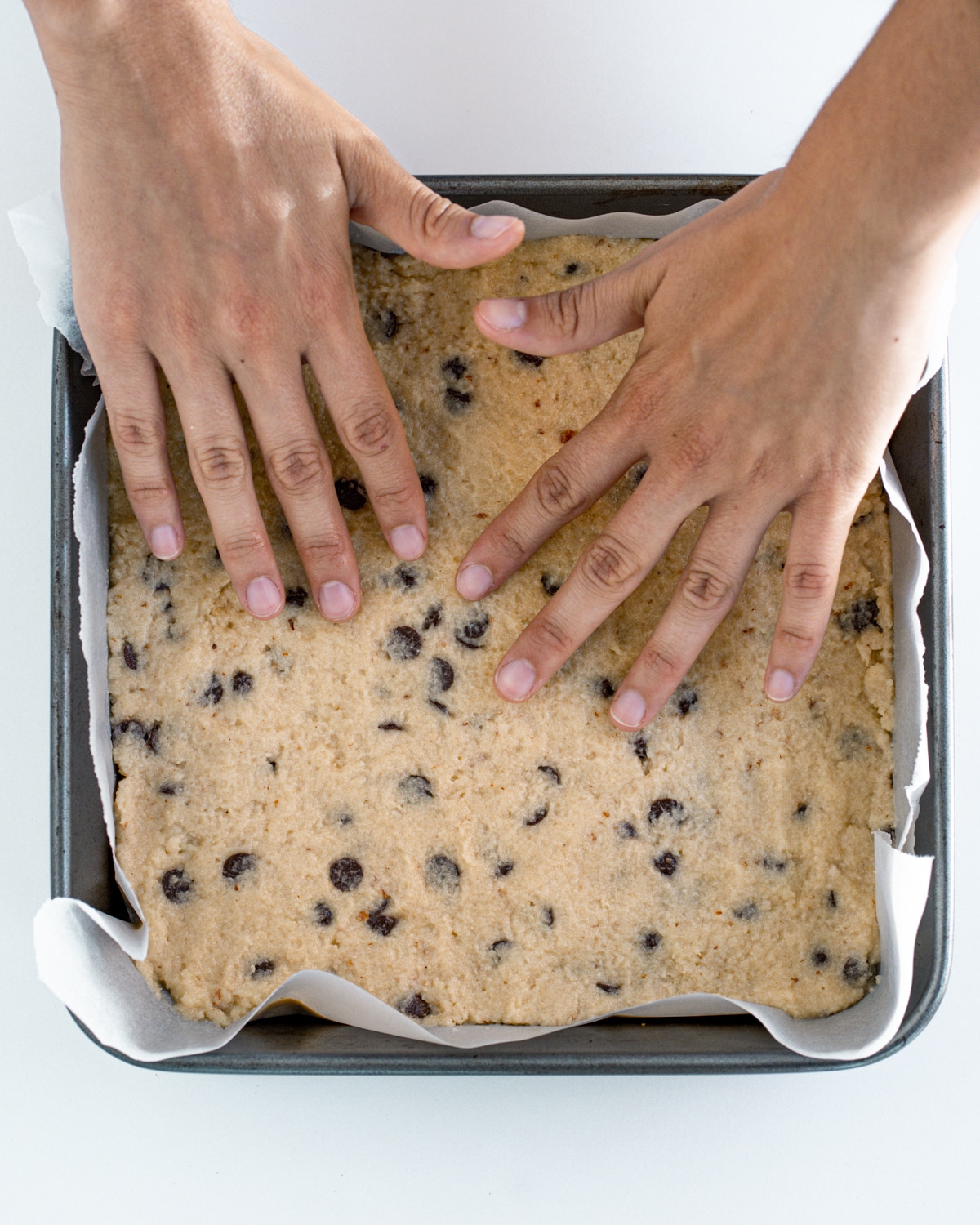 Picture of cookie dough in a square parchment-lined baking dish and hands pressing down on it.