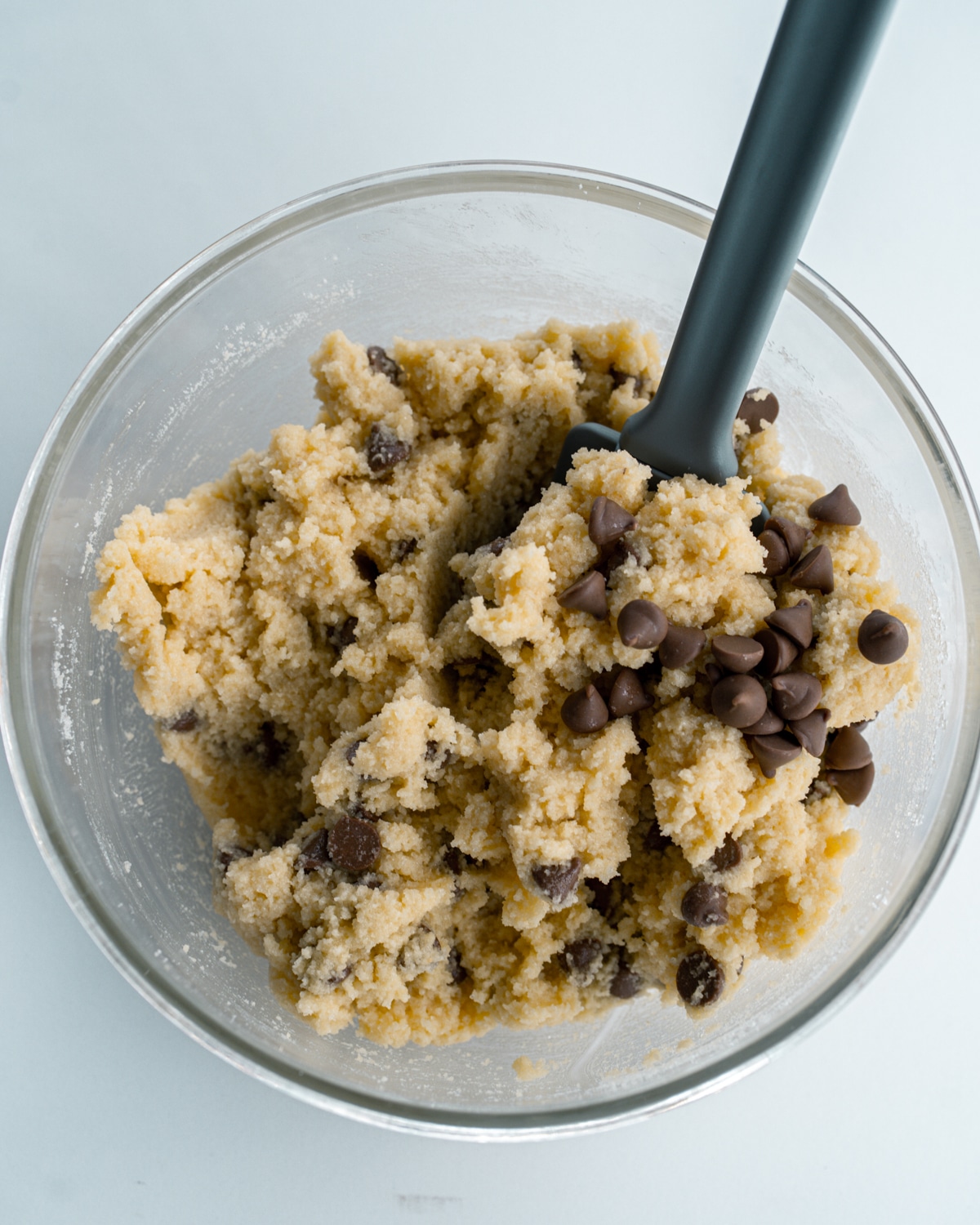 Picture of glass bowl with dough and chocolate chips mixed together and a grey spatula inside.  