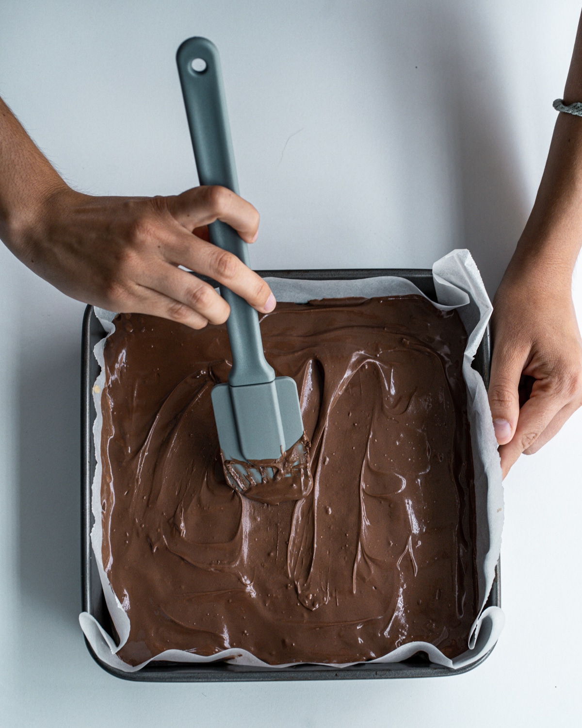 Picture of cookie dough in a parchment lined square baking dish with melted chocolate on top and hands spreading the chocolate with a grey spatula.