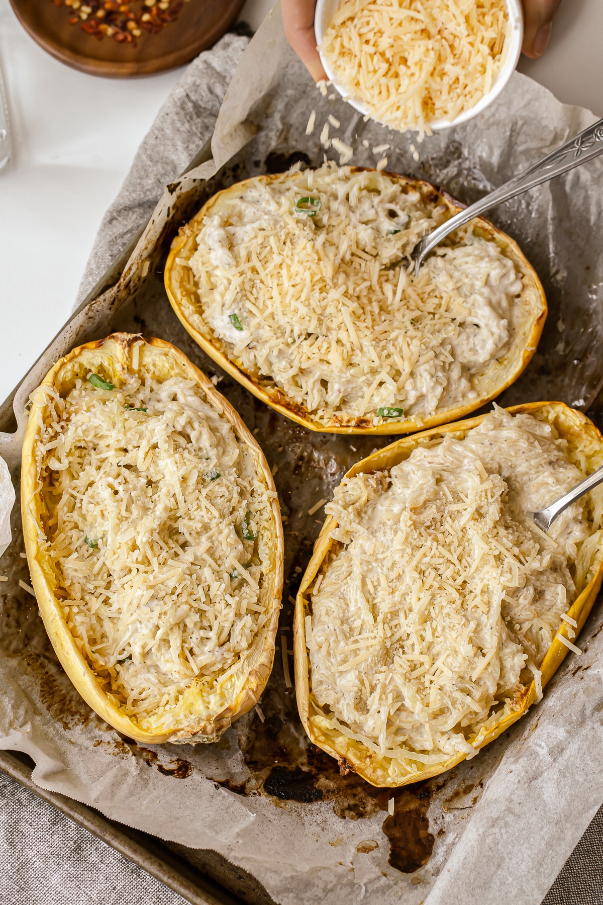 Picture of three halves of Alfredo spaghetti squash with forks in them on a parchment lined baking tray with a hand sprinkling Parmesan cheese on top.