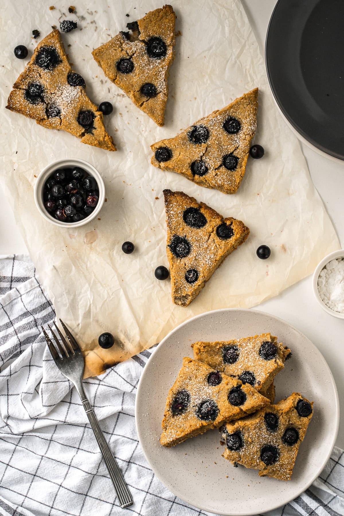 Blueberry scones spread out on a white cloth with a small white jar of blueberries and a fork in the bottom left.