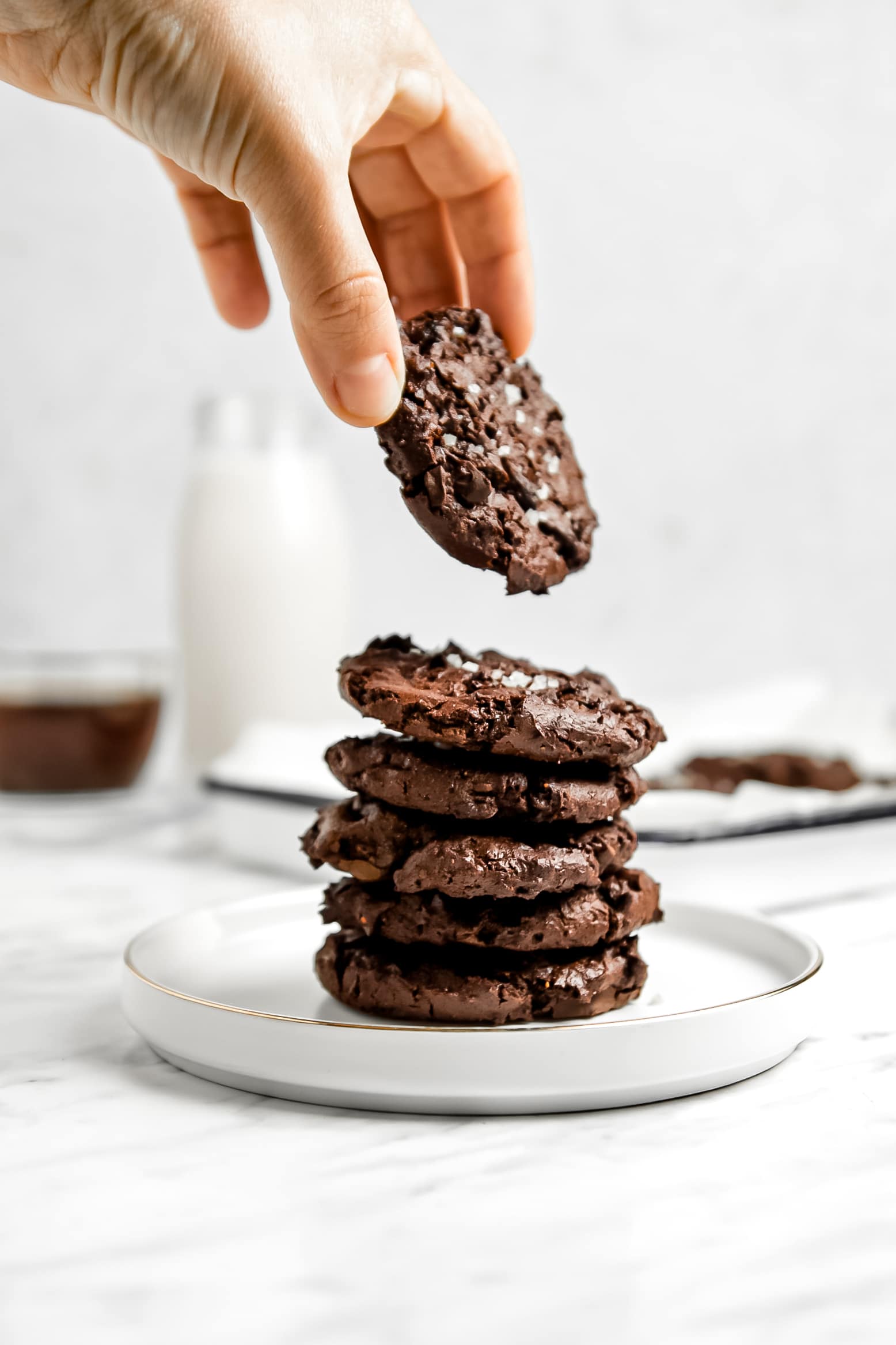 Picture of a stack of chocolate brownie cookies on a small white plate with a hand picking up the cookie on top.