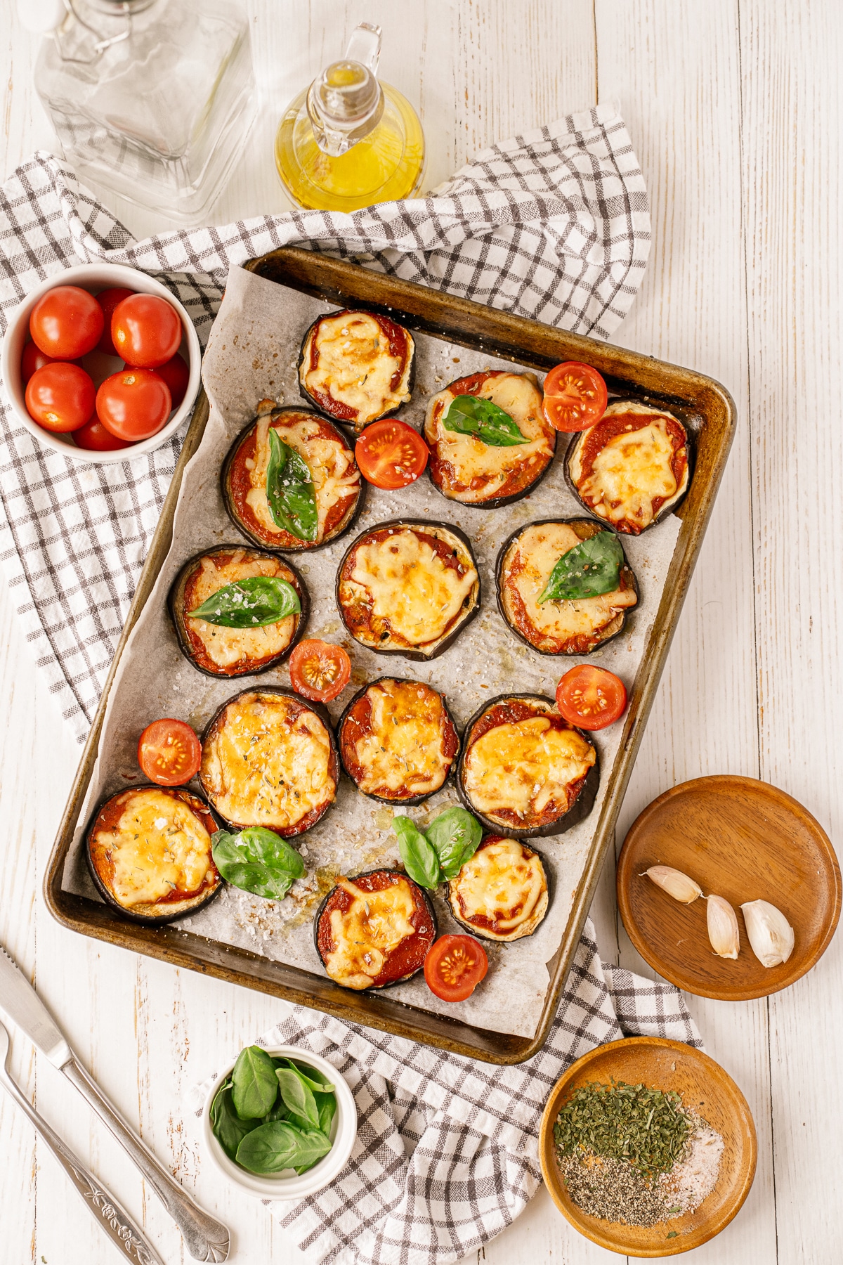 Overhead picture of eggplant parmesan on a baking sheet lined with parchment paper on a checkered towel with a small white bowl of tomatoes in the upper left and a small wooden plate of garlic cloves in the bottom right corner.