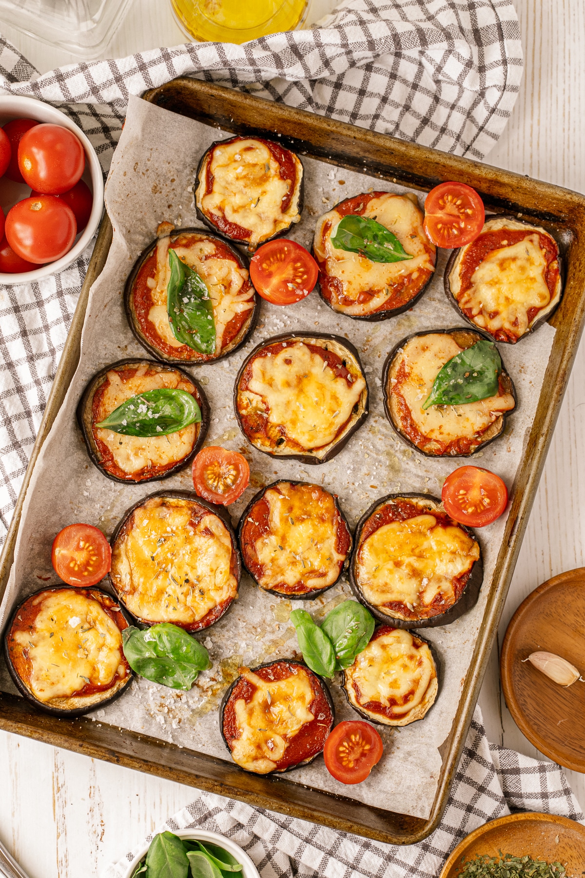 Overhead picture of eggplant parmesan on a baking sheet lined with parchment paper and a small white bowl of tomatoes in upper left corner on a checkered towel.