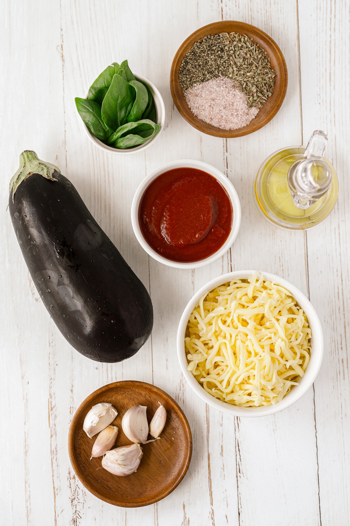 Overhead picture of ingredients for eggplant parmesan on a white background in small white bowls and small wood plates.
