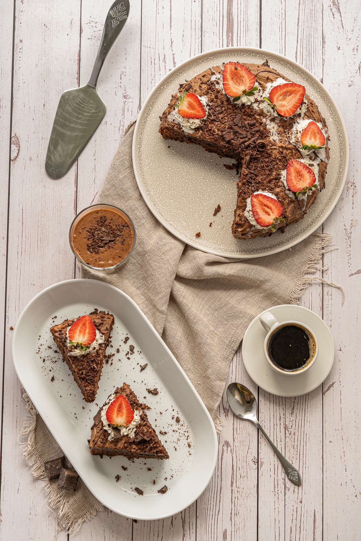 Overhead picture of finished chocolate mousse cake with sliced strawberries on top and two pieces of cake on a white plate to the bottom left and a white mug of coffee to the right.