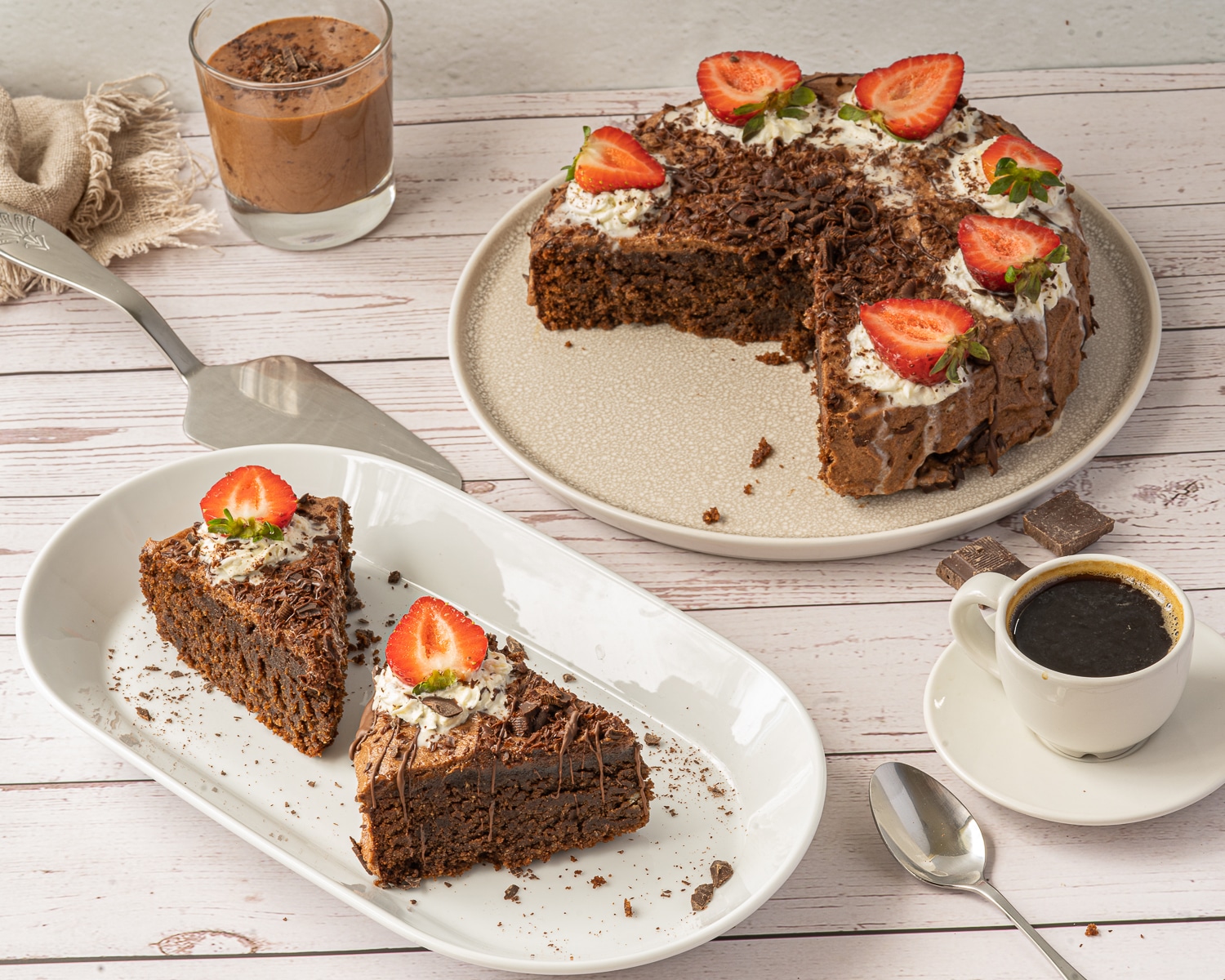 Horizontal picture of finished chocolate mousse cake with sliced strawberries on top and two pieces of cake on a white plate to the bottom left and a white mug of coffee to the right.