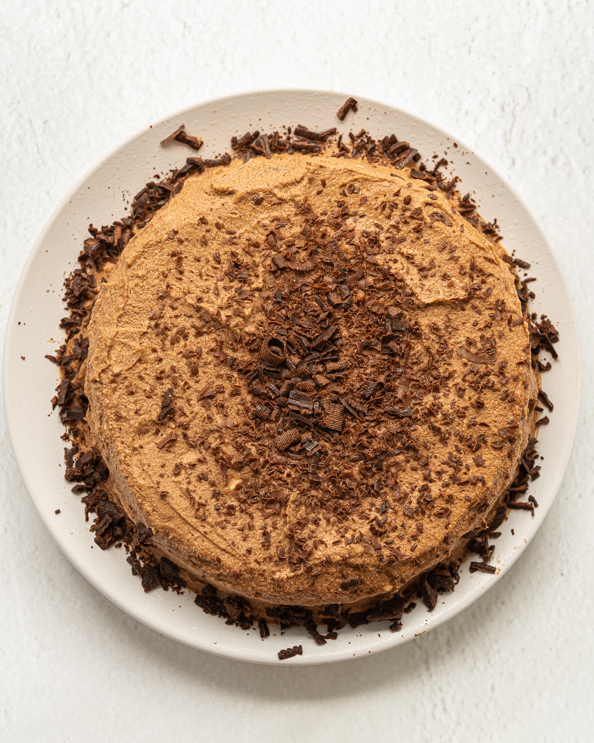 Picture of frosted chocolate mousse cake with chocolate shavings on a white plate on a white background.