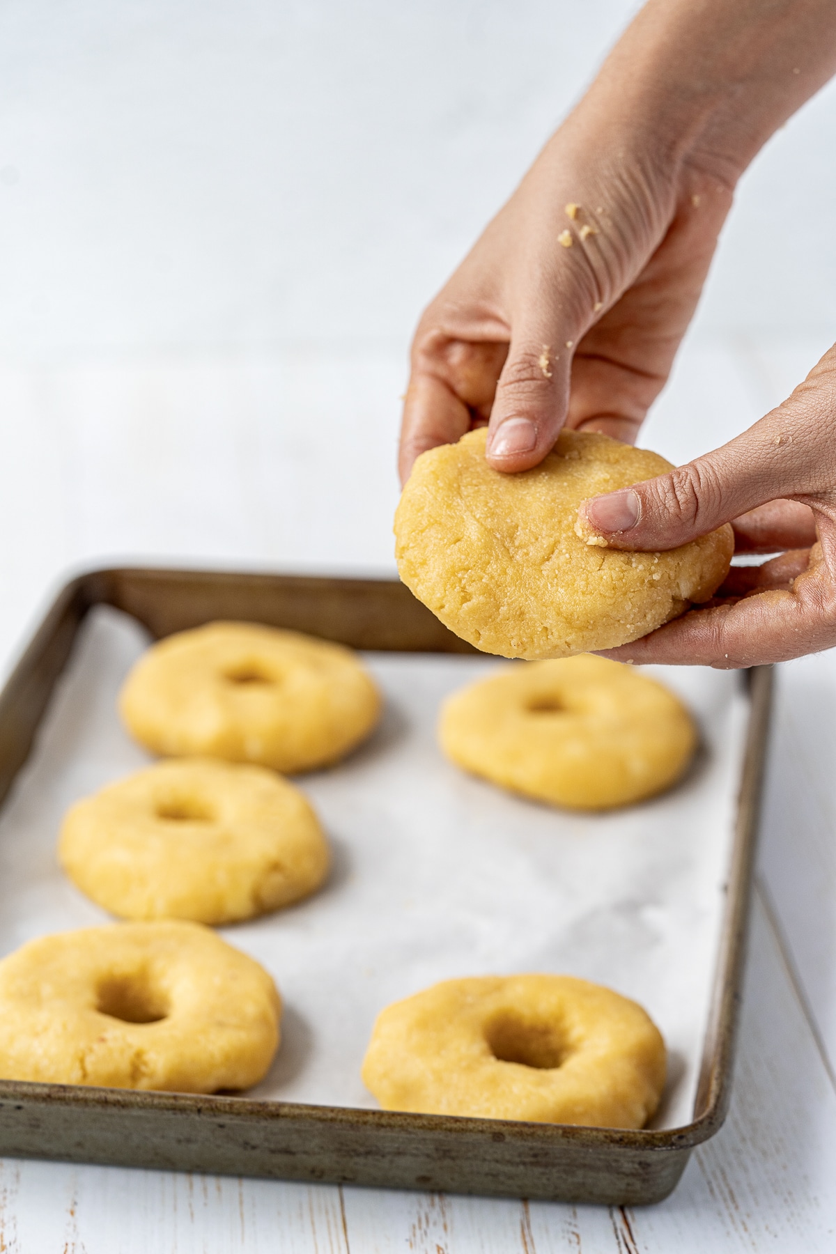 Picture of uncooked low carb bagels on a baking tray lined with parchment paper and a hand holding some dough formed into a circle.
