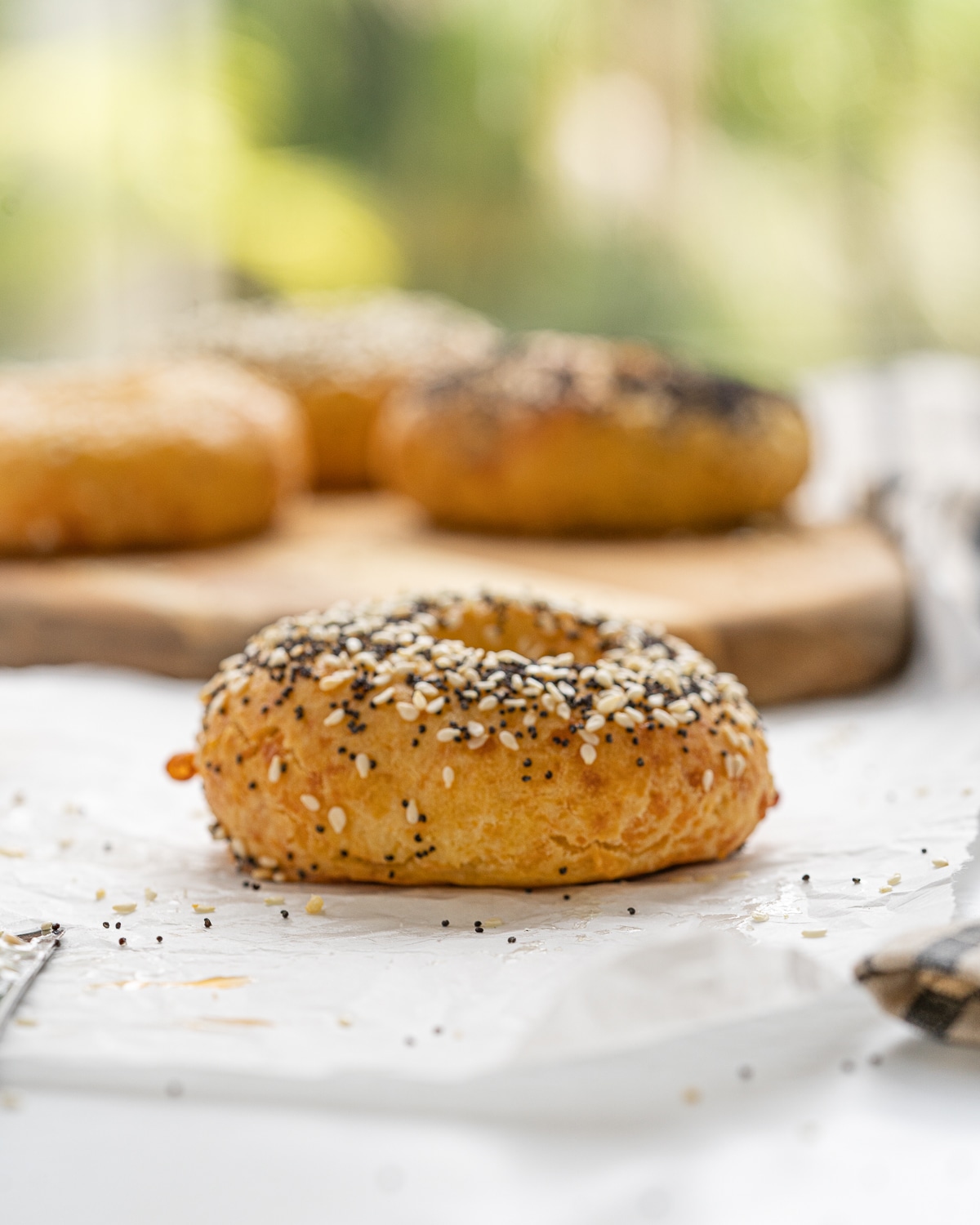 Close up picture of the side of a bagel with everything seasoning on top and its on top of parchment paper with a bunch of bagels on a wooden tray in the background.
