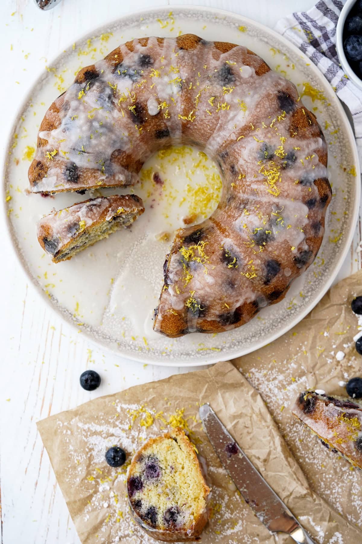 Overhead picture of lemon blueberry cake with icing and lemon zest on top while one a white plate, a slice of the cake on parchment paper in front of the cake.