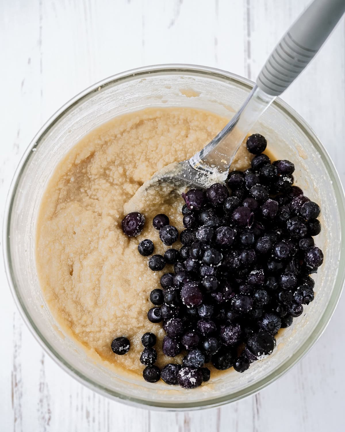 Overhead picture of glass bowl with ingredients mixed together and lemon zest and blueberries on top and a spoon inside.