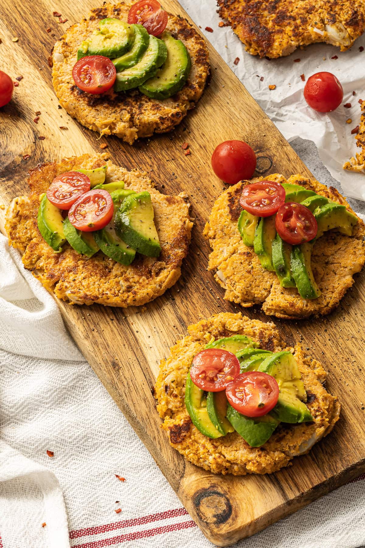 Overhead picture of 4 cauliflower fritters with sliced avocado and tomatoes on top on a long rectangular wood board.