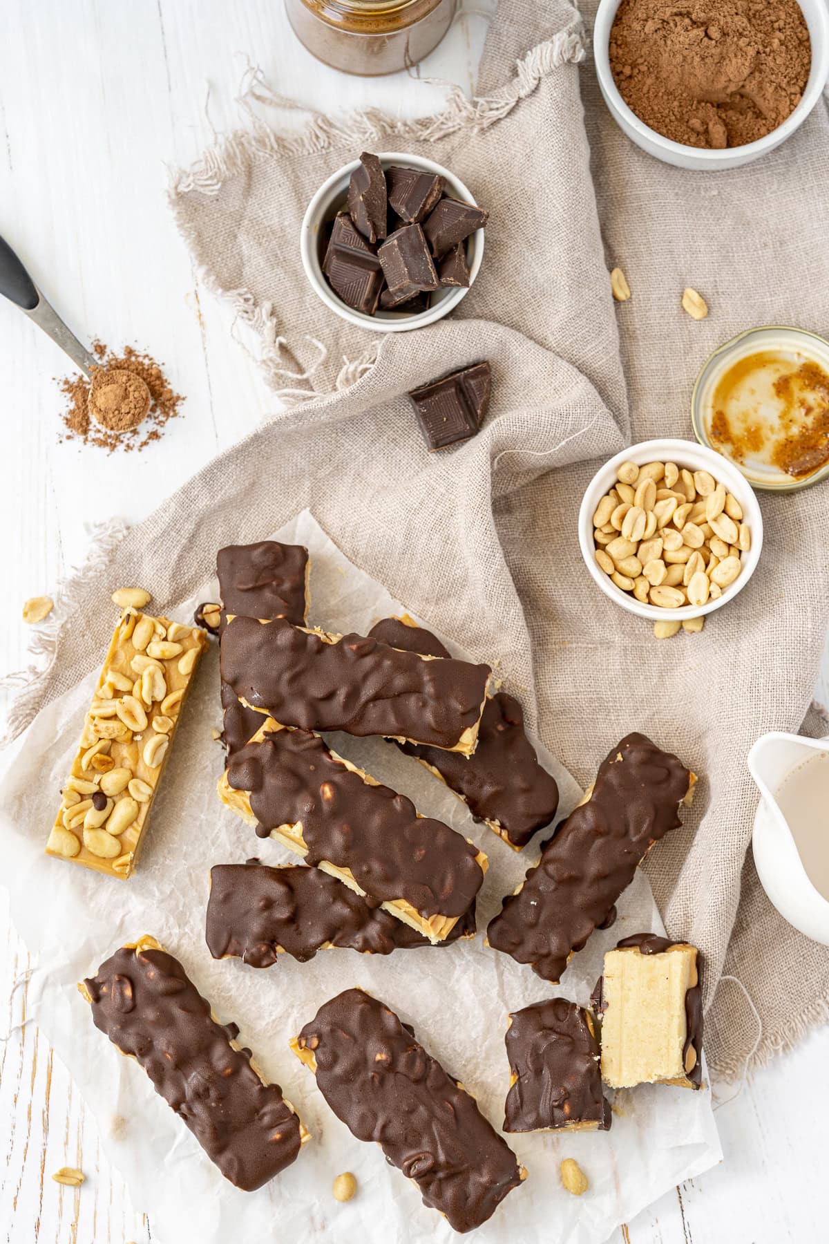 Overhead picture of homemade snickers bars on a beige cloth scattered and piled up with one to the left without chocolate drizzle, a small white bowl of chopped peanuts, a small white bowl of chopped chocolate and a teaspoon full of cocoa powder in the upper left corner.