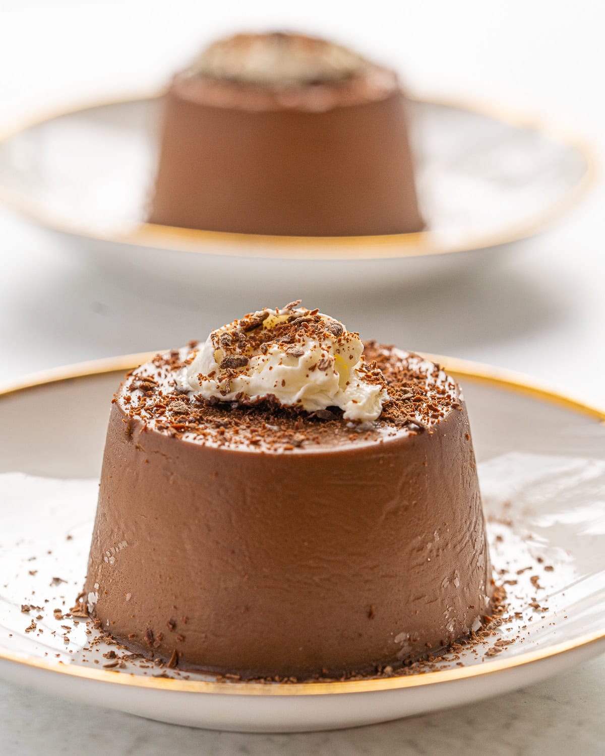 Close up picture of one chocolate Panna Cotta on a white plate with gold trim and whipped cream and shaved chocolate on top and another chocolate Panna Cotta in the background.