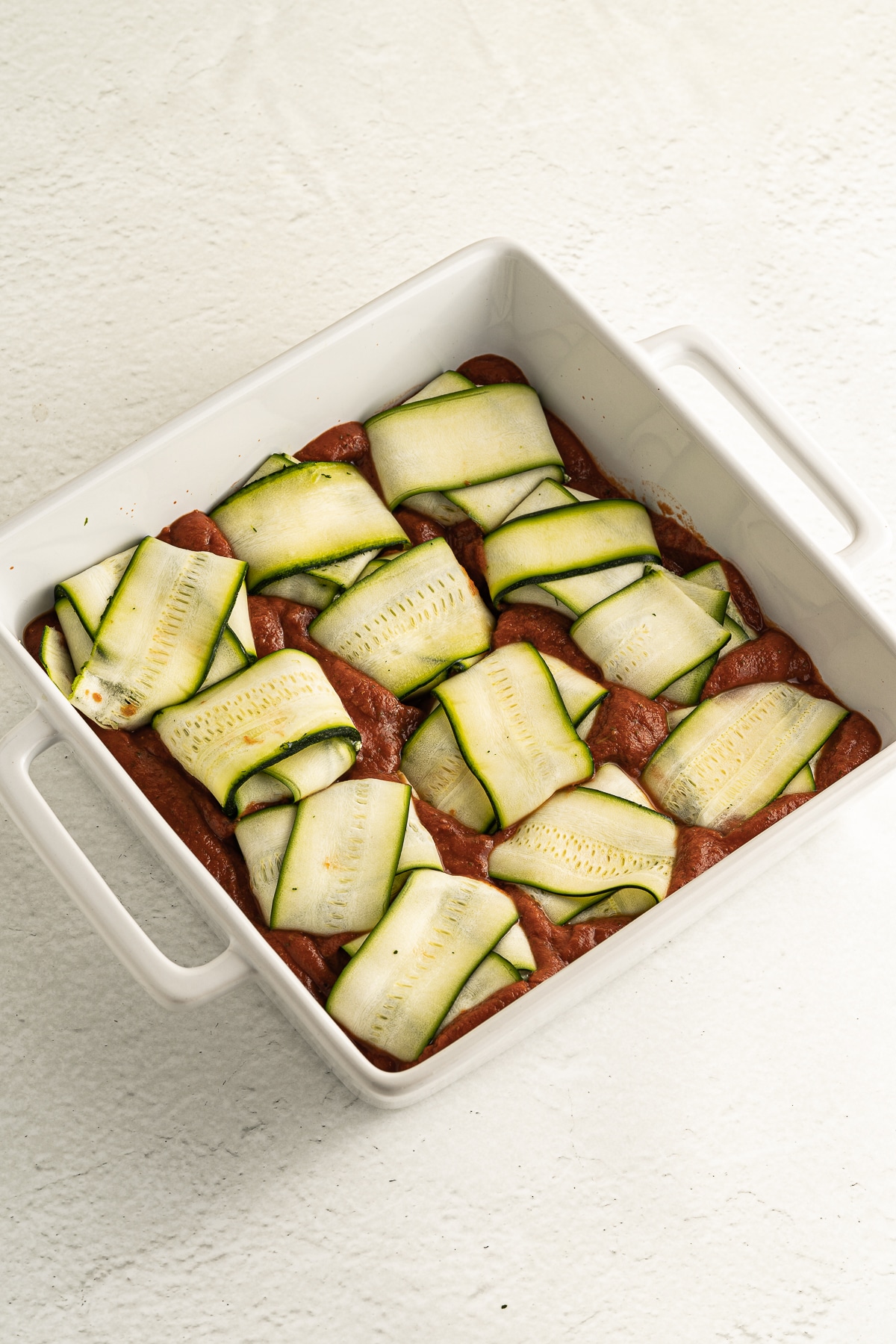Overhead picture of zucchini raviolis laying onto marinara sauce in a white rectangular baking dish.
