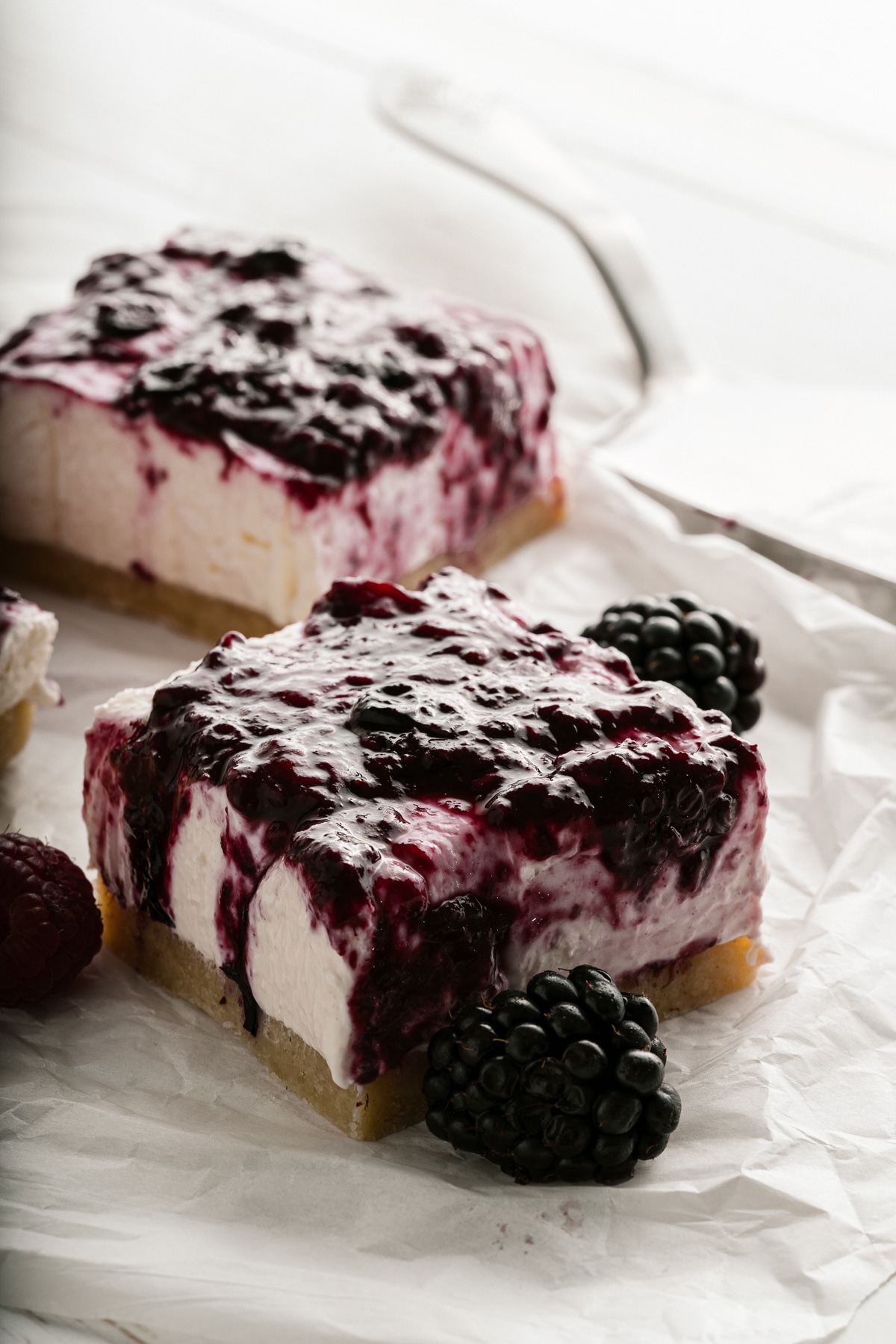 Close up two cheesecake bars on parchment paper with a fresh blackberry in front.