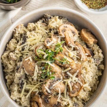 Overhead picture of cauliflower rice risotto in a white bowl on a striped towel with a small white bowl of seasonings in the upper right corner and green spices in the upper left corner.