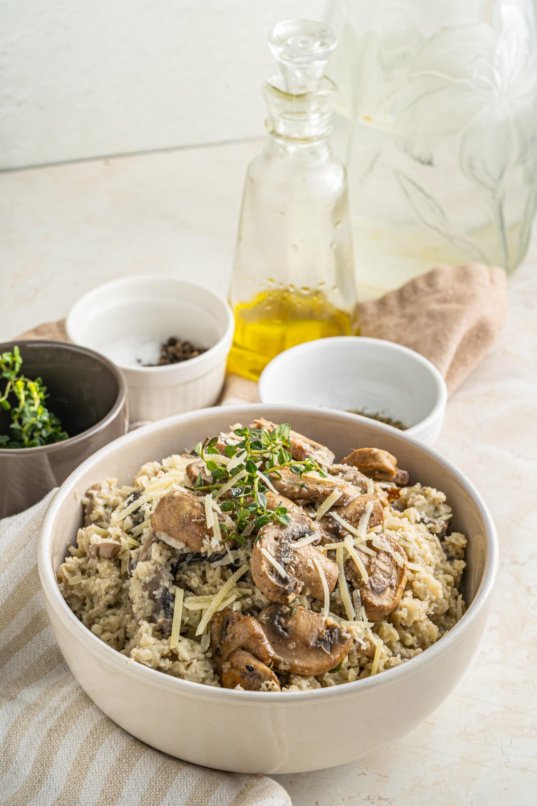 Picture of cauliflower rice risotto close up with mushrooms and shredded cheese on top with two small white jars of seasonings up top behind an olive oil jar and a brown bowl of green spices.
