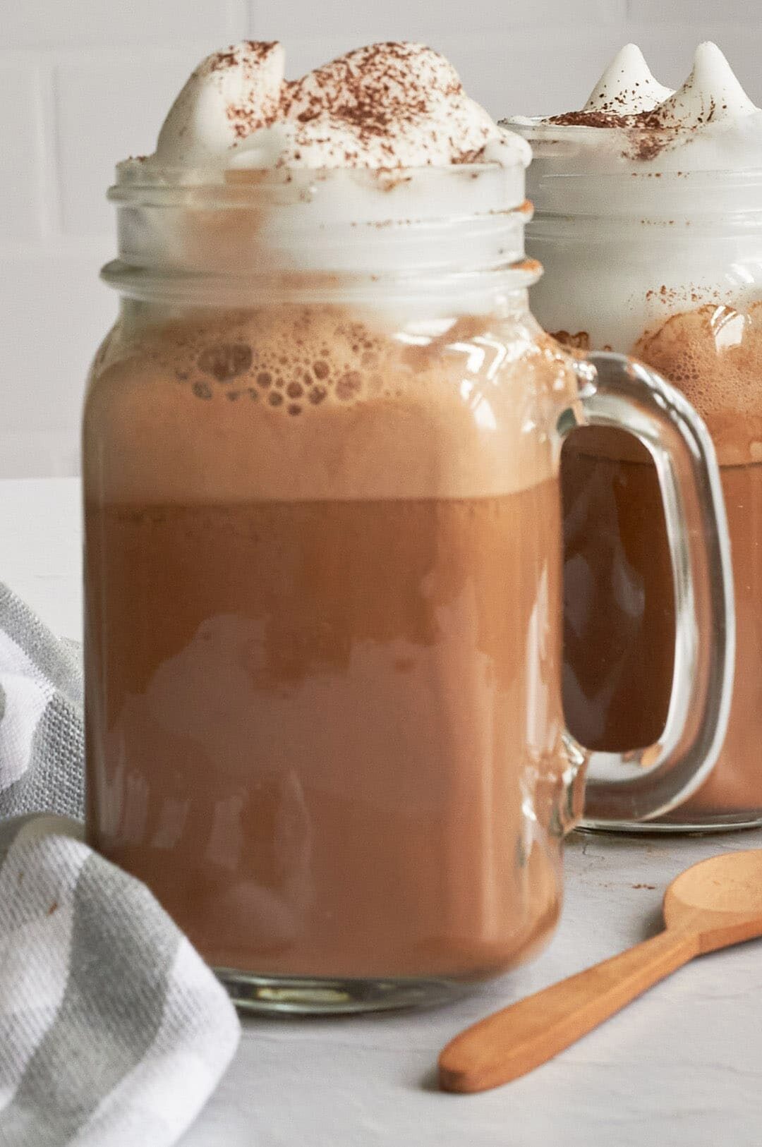 Close up of hot cocoa with whipped cream dripping on top of a clear mason jar mug with a wooden spoon in bottom right corner and a grey and white stripe towel in bottom left corner.