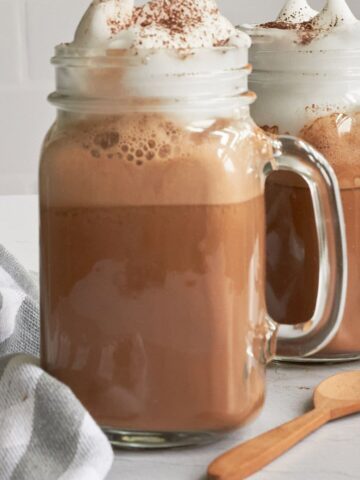 Close up of hot cocoa with whipped cream dripping on top of a clear mason jar mug with a wooden spoon in bottom right corner and a grey and white stripe towel in bottom left corner.