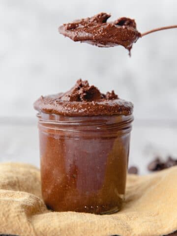 Homemade healthy Nutella recipe in a clear glass jar with a spoonful of Nutella on top on a beige towel.