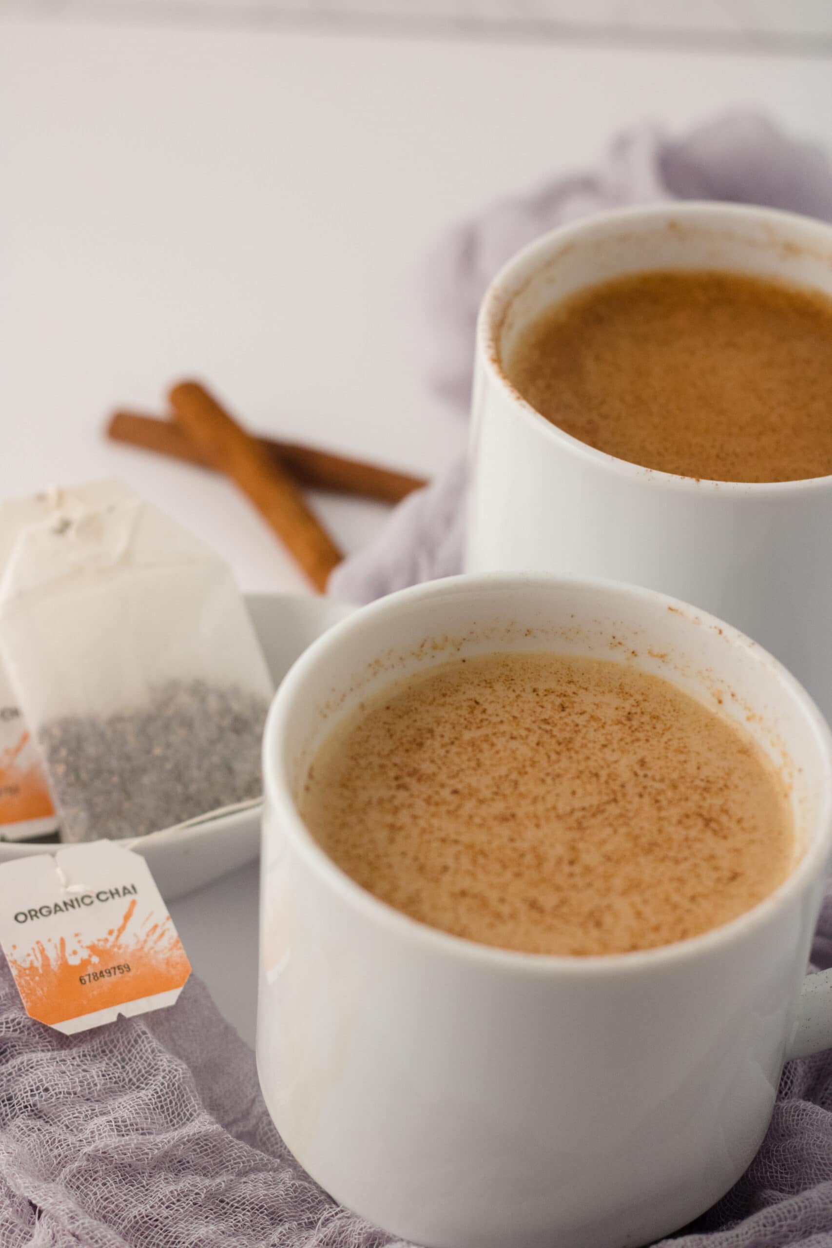 Picture of apple chai latte in a white mug on a grey cloth with tea bags in the corner and another white mug of latte in the back.