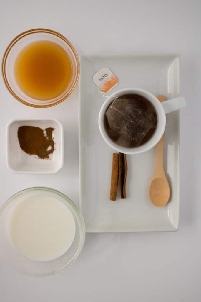 Overhead picture of tea in a white mug on a white plate with a cinnamon stick and wooden spoon and apple cider in upper left corner in a clear glass bowl and cinnamon in a small white bowl and almond milk in a clear glass bowl on bottom left corner.